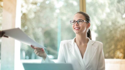 Businesswoman with glasses smiling and holding papers in bright office.